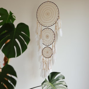 May include: A handcrafted dreamcatcher with three circular woven frames in cream and brown. White feathers hang from the bottom, and the dreamcatcher is suspended against a white wall. The design incorporates intricate crochet details.
