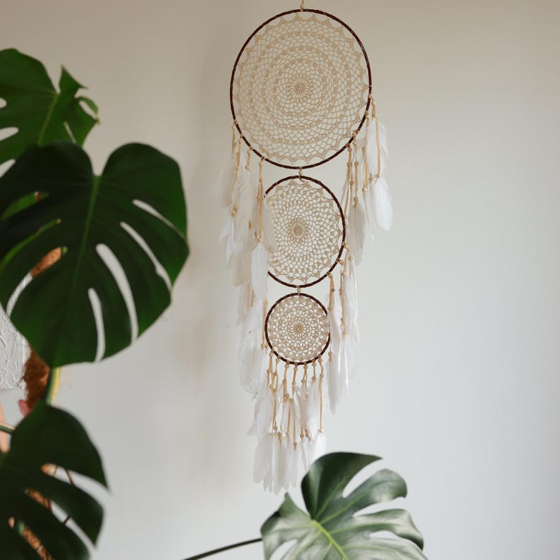 May include: A handcrafted dreamcatcher with three circular woven frames in cream and brown. White feathers hang from the bottom, and the dreamcatcher is suspended against a white wall. The design incorporates intricate crochet details.
