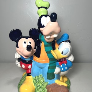 May include: A ceramic cookie jar featuring Mickey Mouse, Goofy, and Donald Duck. Mickey Mouse is wearing red shorts and yellow shoes. Goofy is wearing an orange shirt, blue pants, and a green hat. Donald Duck is wearing a blue sailor suit.