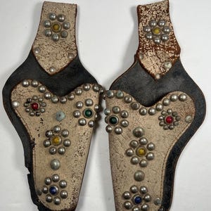 Op de afbeelding: Twee vintage leren pistoolholsters. Elke holster heeft een crèmekleurige voorkant versierd met zilverkleurige studs en kleurrijke edelsteenaccenten. De holsters hebben een zwarte leren achterkant en een verweerde uitstraling.