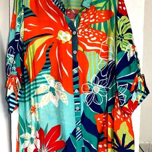 May include: A colourful button-down shirt with a floral design. The shirt features large orange flowers, white and yellow accents, and green leaves against a turquoise and teal background. The sleeves are rolled up and tied. The shirt has a collared neckline.