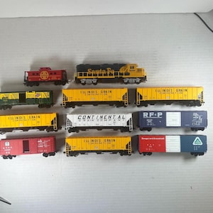 Puede incluir: Una colección de once vagones de tren a escala que incluye una locomotora Santa Fe amarilla y negra, un furgón de cola rojo y varios vagones de carga con las etiquetas Illinois Grain Corporation, Continental Grain Company, RF&P y Bangor and Aroostook.