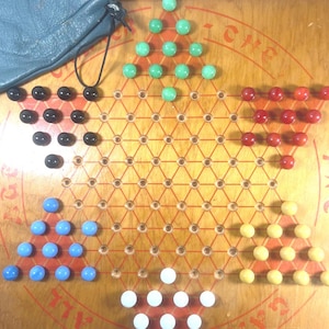 May include: Wooden Chinese Checkers game board with red lines and holes. The game pieces are various colors: green, black, red, blue, yellow, and white. A blue leather drawstring bag is in the upper left corner. The game is labeled "THE GAME OF ALL GAMES".