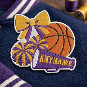 May include: Embroidered patch featuring a basketball, bow, pom-poms, and megaphone with "ANYNAME" text. The patch has a white border, with gold, purple, and orange colors. The patch is on a navy blue jacket.