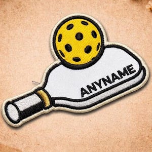 May include: Embroidered patch featuring a yellow pickleball ball with black dots atop a white paddle. The paddle has black and gold accents and the word "ANYNAME" in black. The patch is outlined in black.