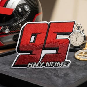 May include: A red and black embroidered patch with the number 95 and the text "ANY NAME" in white. The patch is on a dark gray surface, with a racing helmet in the background. A stopwatch and a gold medal are also visible.