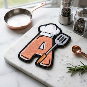 May include: Embroidered patch in the shape of the letter "A" with a chef's hat and spatula design. The patch is peach-colored with black trim. A copper-colored measuring spoon, a small pan, and a pepper grinder are also visible.