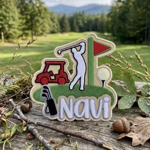 May include: Embroidered patch featuring a golf theme. The design includes a golfer, golf cart, golf ball, and flag. The name "Navi" is embroidered in white. The patch is set against a green and beige background, with a wooden surface and natural elements.