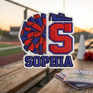 May include: Embroidered patch with a red, white, and blue pom-pom design and the letter "S". The name "Sophia" is embroidered below. A varsity game program is visible.