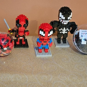 May include: A collection of block figures featuring Marvel characters Deadpool, Spider-Man, and Venom. The figures are red, black, and white, and are displayed on small grey and black bases. Two clear plastic baubles contain additional block pieces.