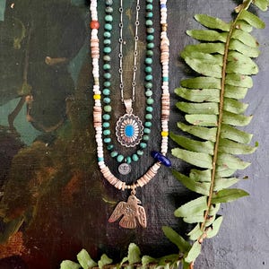 May include: A collection of necklaces with turquoise, white, and brown beads. One necklace features a silver pendant with a turquoise stone and a sunburst design. Another has a gold-colored bird pendant. The necklaces are displayed on a dark, textured background with a fern.