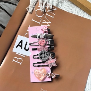 May include: A collection of hair clips in various designs, including stars, hearts, and a smiley face. The clips are silver-toned with pink and clear accents. The clips are displayed on a pink card.
