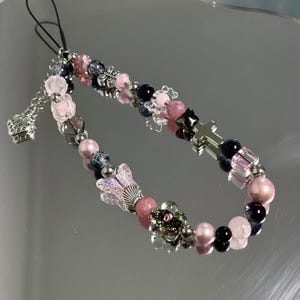 May include: A beaded phone charm featuring pink, black, and silver beads. The charm includes a silver cross, butterfly, and dice charm. The beads are various shapes and sizes, creating a unique accessory.