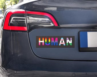 May include: A rectangular bumper sticker applied to the trunk of a dark gray car. The sticker features the word HUMAN in bold capital letters, with each letter filled with a different pride flag pattern, including rainbow, bisexual, and transgender colors.