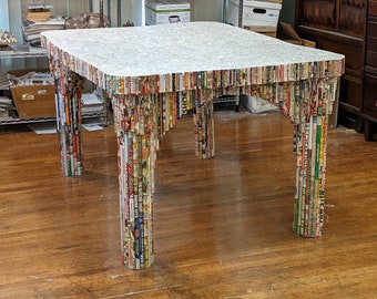 May include: A square table with a white top and four legs made of rolled up magazines. The legs are made of a variety of magazines with different colors and patterns.