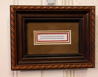 May include: A framed quote in a dark brown frame with a gold-colored border. The quote reads: "The early bird gets the worm, but the second mouse gets the cheese."