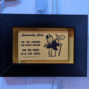 May include: Framed vintage Monopoly Community Chest card. The card features text that reads "YOU ARE ASSESSED FOR STREET REPAIRS" and "$40 PER HOUSE, $115 PER HOTEL." The card is set against a yellow background within a black frame.