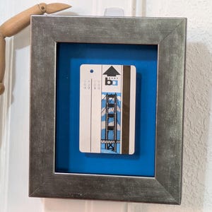 May include: A rectangular silver-toned frame containing a white transit card with a graphic illustration of the Golden Gate Bridge and the text BART. The card is mounted against a solid blue background, creating a shadow box effect.