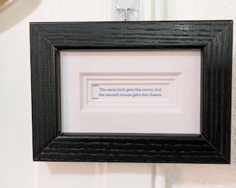 May include: A rectangular black wooden frame containing a white mat with a recessed center. The white paper insert features the blue printed text: The early bird gets the worm, but the second mouse gets the cheese. The frame has a visible wood grain.