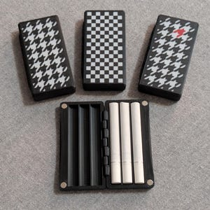 Puede incluir: Un estuche de cigarrillos negro abierto con tres cigarrillos blancos, rodeado de tres estuches de cigarrillos con diseños en blanco y negro. Los diseños incluyen pata de gallo y un diseño de tablero de ajedrez. Un estuche tiene un corazón rojo.