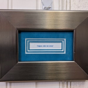 May include: A silver framed picture with a blue background and a white card inside that says "Calgon, take me away!"