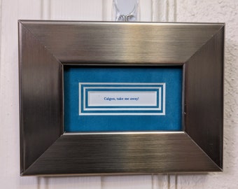 May include: A silver framed picture with a blue background and a white card inside that says "Calgon, take me away!"