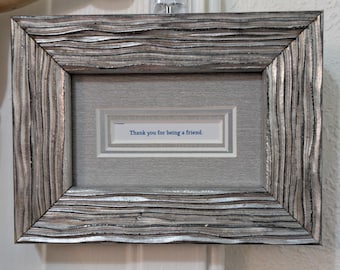 May include: A rectangular picture frame with a textured, wavy silver and grey finish. The frame contains a light grey mat with a central white insert featuring the blue printed text, Thank you for being a friend. The design is clean and structured.