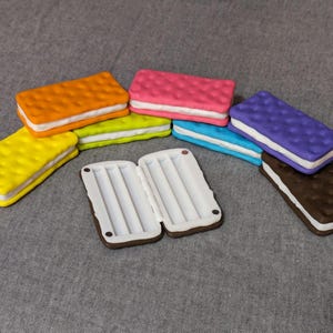 May include: A collection of colorful, toy ice cream sandwiches in various colors, including orange, pink, yellow, green, blue, purple, and brown. Each sandwich has a white filling and a textured surface.