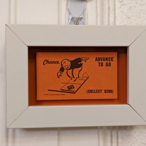 May include: A framed Monopoly Chance card with an orange background. The card shows a cartoon man in a top hat and monocle running towards the 'Go' space on the board. The text on the card reads "Advance to Go (Collect $200)".