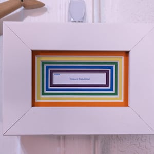 May include: A white framed art piece with a colorful layered design. The artwork features a rectangular frame with orange, yellow, green, blue, and purple layers. The text "You are Sunshine!" is visible within the inner frame.