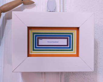 May include: A white framed art piece with a colorful layered design. The artwork features a rectangular frame with orange, yellow, green, blue, and purple layers. The text "You are Sunshine!" is visible within the inner frame.