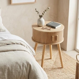 Op de afbeelding: Een licht houten nachtkastje met een ronde bovenkant en drie schuine poten. De tafel heeft een geribbeld ontwerp op de lade en een kleine houten handgreep. Een vaas, boek en bril staan erop. Een ingelijste botanische print hangt aan de muur.