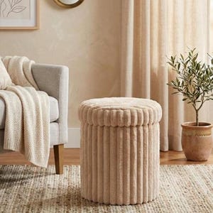 Op de afbeelding: Een beige, ronde poef met een geribbelde, pluche buitenkant. De cilindrische poef staat op een geweven vloerkleed. Een lichtgekleurde bank en een plant in een pot staan op de achtergrond.
