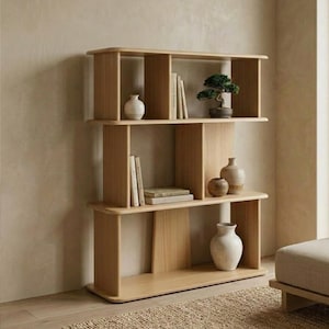 Puede incluir: Una estantería de madera clara con tres estantes. Los estantes exhiben libros, pequeños jarrones y un bonsái. La estantería tiene un diseño moderno con bordes redondeados y soportes verticales.