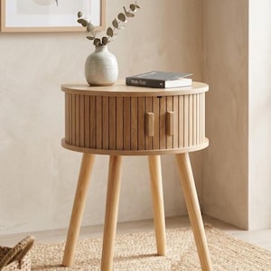 May include: A light wood, round side table with three angled legs. The table has a ribbed, cylindrical body with two small doors. A small vase with greenery and a book are on top. The table is 24 inches tall.