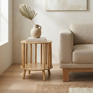 Può includere: Un tavolino rotondo in legno chiaro con supporti verticali e un ripiano inferiore. Sopra poggia un vaso in ceramica beige opaco contenente una foglia di palma essiccata, posizionato su due libri bianchi accanto a un divano beige imbottito.