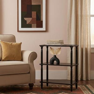 Puede incluir: Escena de salón con una obra de arte abstracta enmarcada con formas geométricas marrones y verdes. Una mesa auxiliar negra de tres niveles con libros y un jarrón decorativo con hojas secas. Un sillón beige con un cojín dorado está cerca.