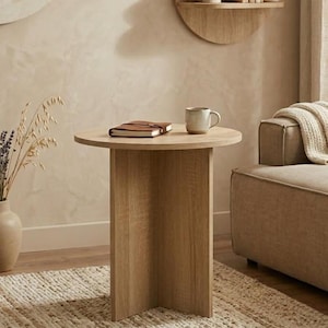 Może przedstawiać: Jasnobrązowy, okrągły stolik boczny z pojedynczą podstawą. Na nim brązowy notatnik w skórzanej oprawie i ceramiczny kubek. Stół stoi na tkanej macie, z beżową sofą i dekoracją ścienną w tle.