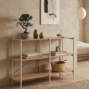 May include: A three-tiered light wood shelving unit with a thin white metal frame. The shelves hold a potted bonsai tree, two ceramic vases, a small metal object, two stacks of books, a stone sculpture, and a woven wicker basket on the bottom shelf.
