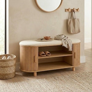 May include: A light wood entryway bench with a cushioned top, two shelves, and two doors. The bench has a curved design and is paired with a round mirror, a woven basket, and a beige tote bag hanging on the wall.