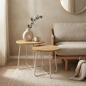 Könnte beinhalten: Zwei runde Beistelltische mit hellen Holzplatten und cremefarbenen Metallgestellen. Auf dem höheren Tisch stehen eine beige Keramikvase mit einer kreisförmigen Öffnung und eine kleine beige Tasse. Die Tische haben ein Kreuzgestell.