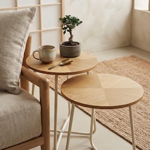 Può includere: Un set di due tavolini rotondi impilabili con piani in legno di rovere chiaro e sottili gambe in metallo bianco. Un piccolo bonsai verde in un vaso grigio, una tazza in ceramica maculata e un cucchiaino in metallo dorato poggiano sul tavolo.