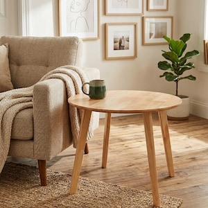 Pode incluir: Uma pequena mesa lateral redonda de madeira clara com três pernas anguladas. Uma caneca verde está sobre a mesa. Uma poltrona bege com um cobertor e uma pequena planta em vaso estão no fundo. Arte emoldurada decorativa na parede.
