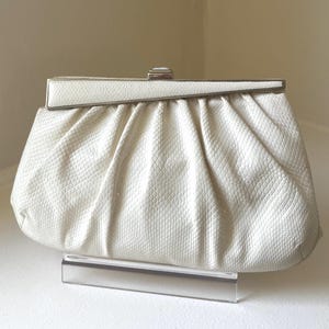 May include: A white clutch bag with a textured snakeskin pattern. The bag features a gathered, pleated design along the top and a silver-toned metal frame closure with a rectangular clasp centered at the top edge of the structured frame.