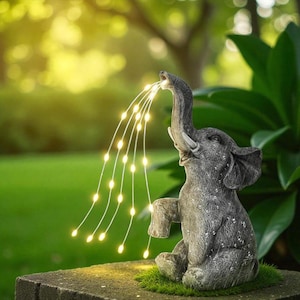 May include: A gray elephant garden statue with a trunk raised, appearing to spray a stream of small, warm white lights. The elephant is seated on a stone surface with green moss, set against a blurred green background.