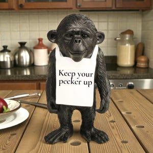 May include: A black chimpanzee figurine wearing a white apron with the text "Keep your pecker up". The figurine is standing on a wooden table. Kitchenware is visible in the background.