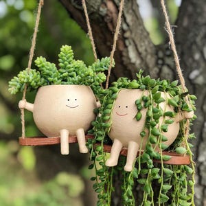 Puede incluir: Dos macetas beige sonrientes, con forma de dibujos animados, con brazos y piernas, cada una sosteniendo un columpio de madera. Una maceta tiene suculentas verdes erguidas, y la otra tiene plantas verdes colgantes. Las macetas cuelgan de cuerdas.