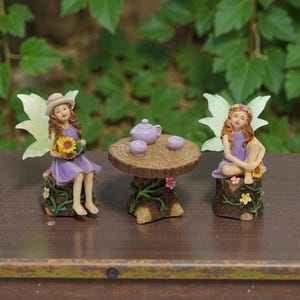 Puede incluir: Tres figuras de hadas. Una hada sostiene girasoles y lleva un vestido morado y un sombrero. Otra hada está sentada y lleva un vestido morado. Una pequeña mesa de madera con una tetera y dos tazas está entre ellas.