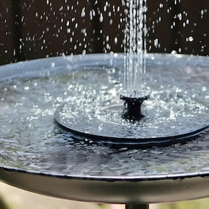 Puede incluir: Una pequeña fuente oscura con agua cayendo en cascada desde una boquilla central. La fuente es redonda, con una cuenca poco profunda llena de agua. Gotas de agua suspendidas en el aire, creando un efecto visual refrescante.