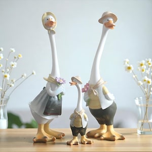 Può includere: Tre statuette decorative di oche. L'oca più alta indossa un cappello e un gilet, l'oca centrale è vestita con un abito e un cappello, e l'oca più piccola indossa un gilet e pantaloncini. Tutti tengono dei fiori.